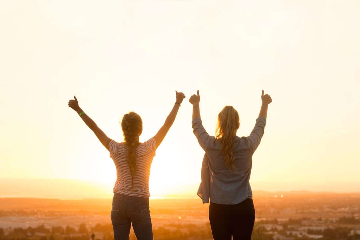 Two women holding their arms up in the air and giving a thumbs up during a bright sunrise.