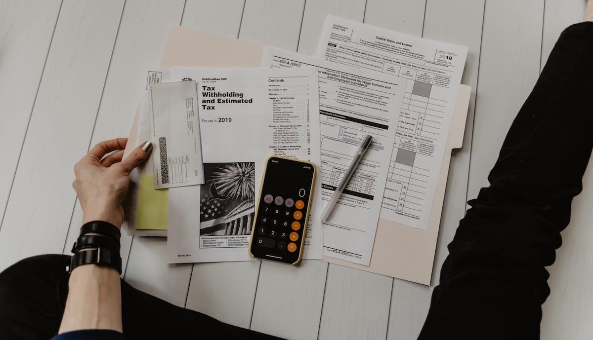 A person’s view of tax papers on the floor and a phone opened to the calculator app. 
