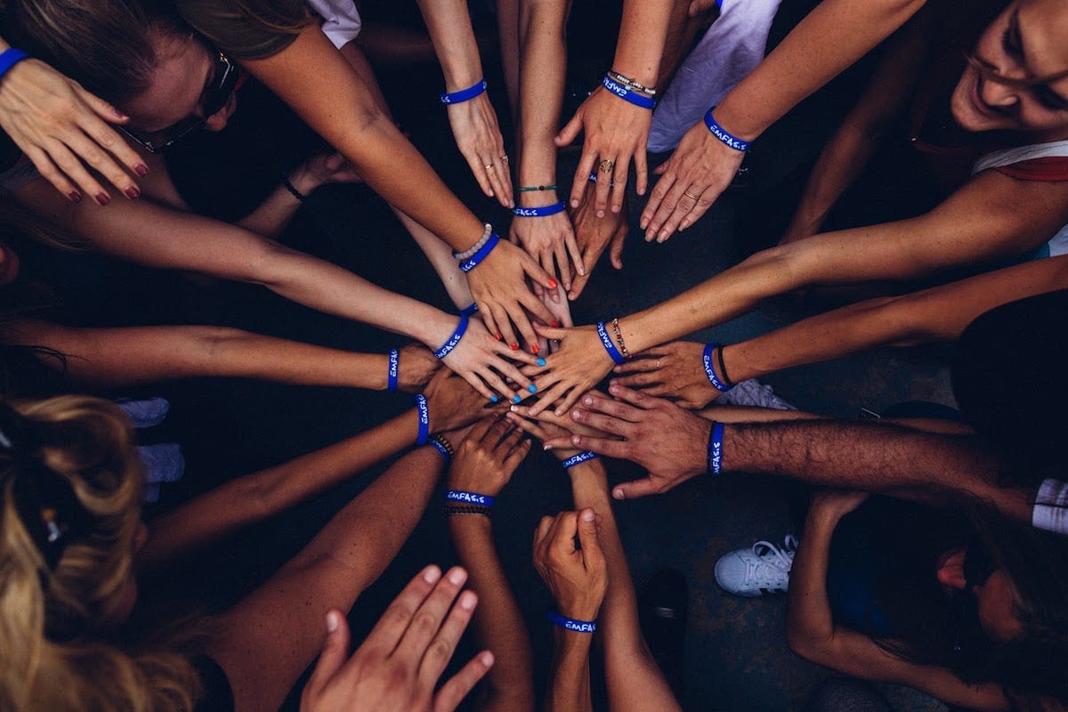 An overhead view of a group of students standing in a circle with one hand per person touching in the center.