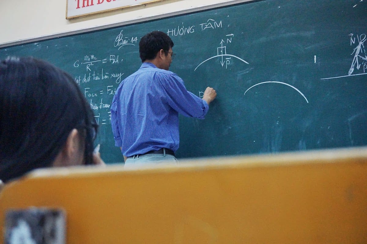 A teacher writes on a chalkboard with their back facing a student