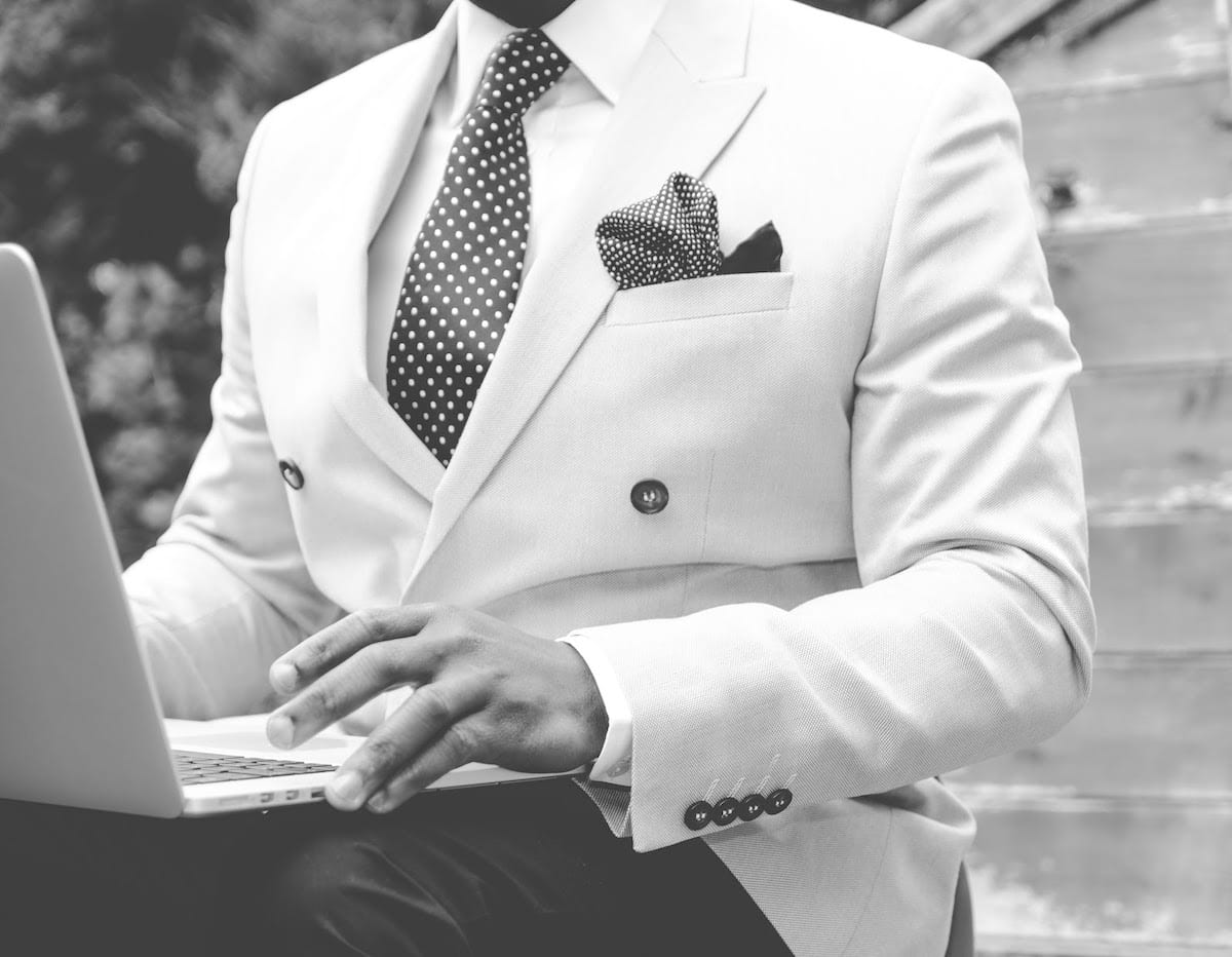 A black and white image of a man wearing a white suit jacket, sitting outside with an open laptop on his lap.