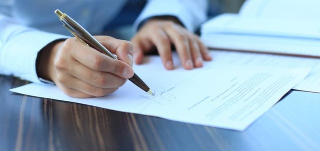 A hand holding a black pen signing a contract on a dark wooden table.