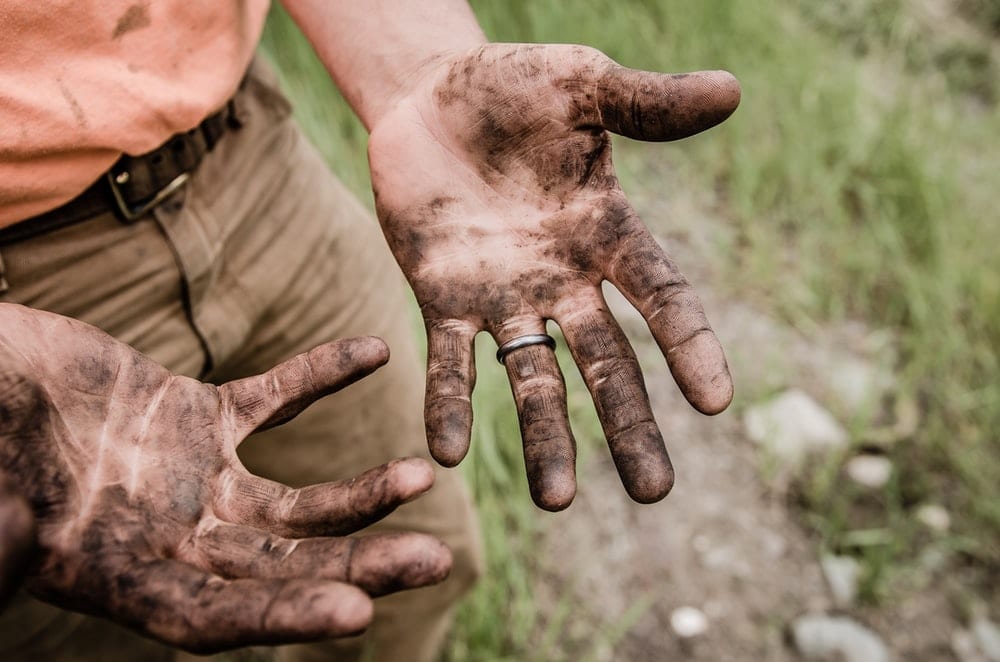 A partial view of a person standing in a field with open, dirty hands.