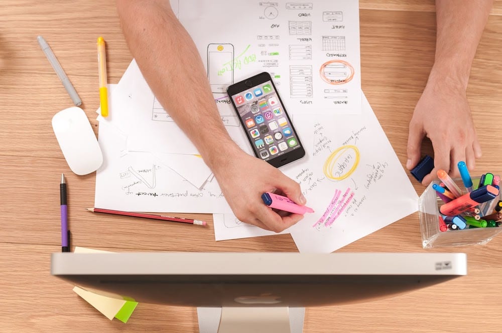 Person using pink highlighter on messy pile of white printer paper in front of computer screen