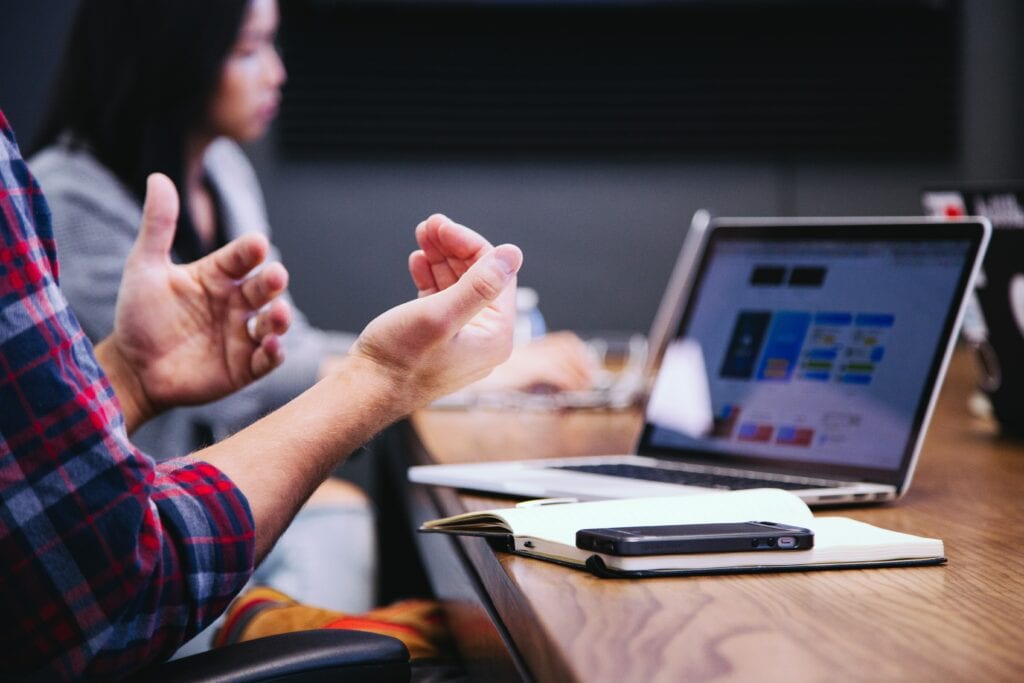 A close-up image of a pair of hands dictating speech with a laptop and a woman in the background.