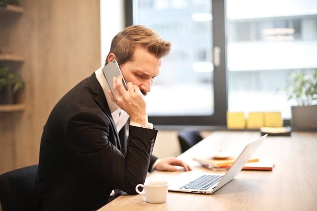 A man sitting in an office setting with a laptop and mug in front of him, conducting a technical screening interview on the phone.