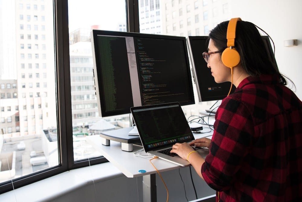 A programmer sitting at a desk by a window with multiple computer monitors, all showing lines of code.