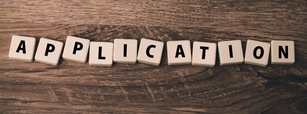 Letter tiles arranged to spell the word application, laid out on a brown desk.
