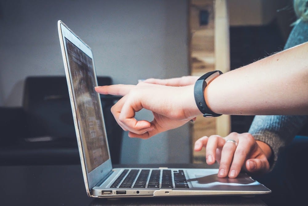 An open laptop with one hand on the trackpad and one hand pointing at the screen.