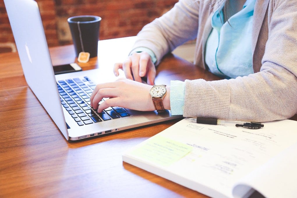 A person writing on a laptop with an open book.