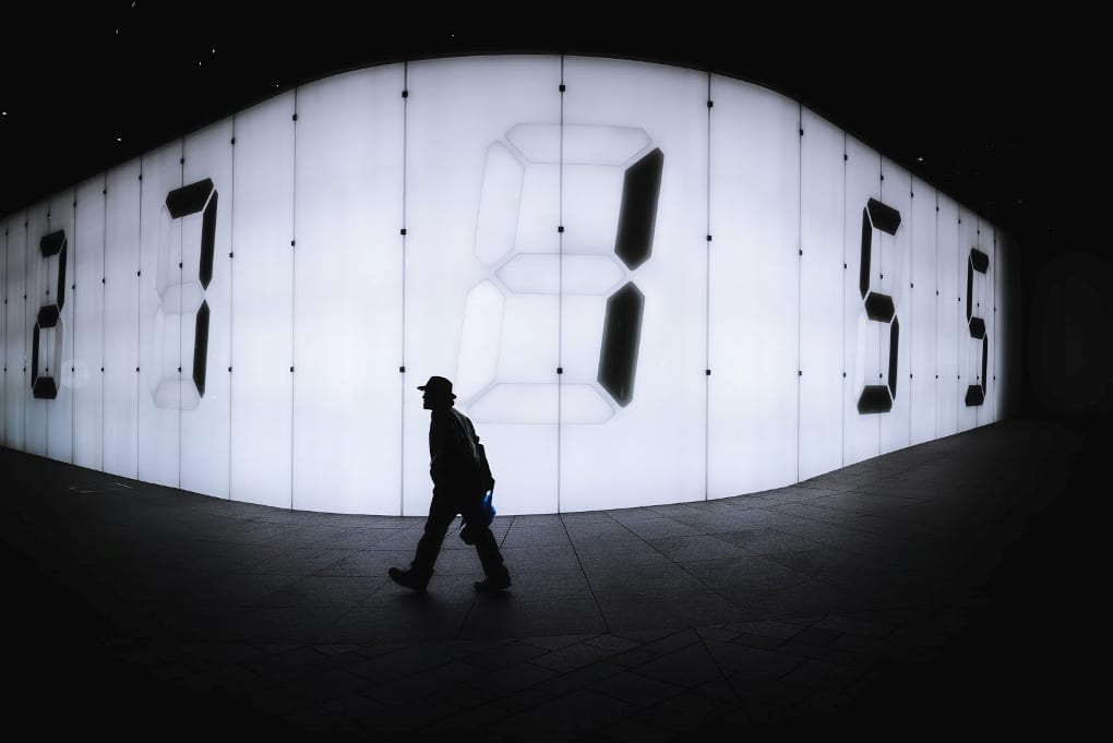 Silhouette of a man walking in front of an illuminated wall with clock numbers.
