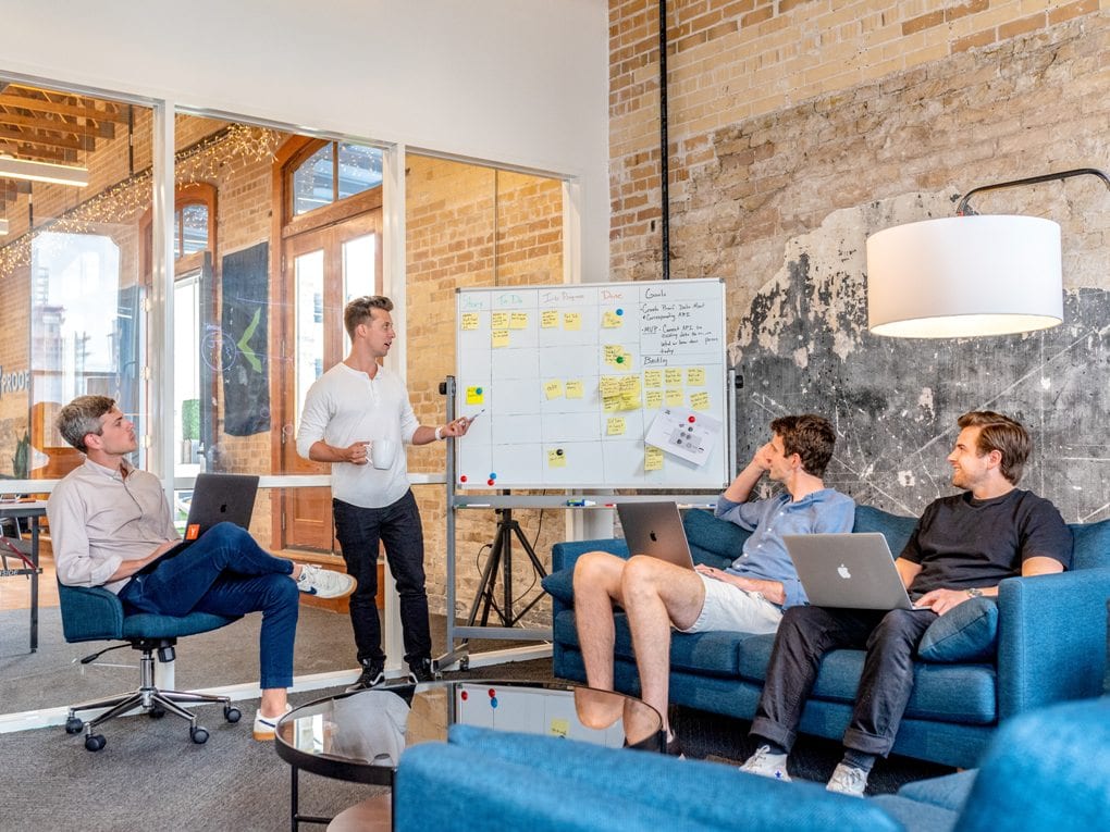 In a conference room, one man standing at a white board covered in post-it notes explains something to three men on laptops seated around him.