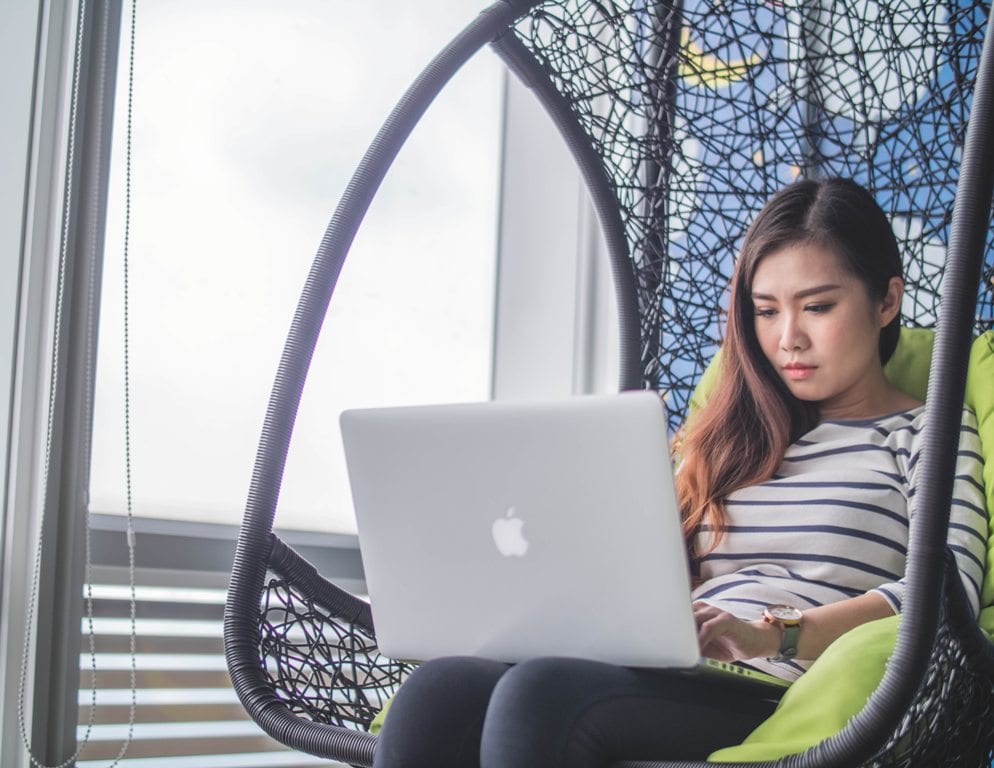 A girl sitting in a wicker chair with a green cushion while working on her computer near a window.
