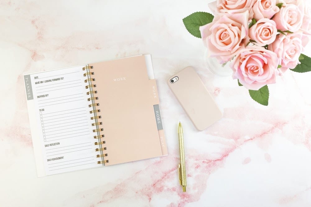 A planner, pen, mobile phone, and pink roses in a vase sit atop a patterned surface