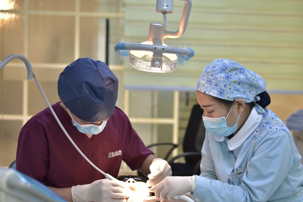 A dentist performs a procedure on a patient while a dental assistant holds a suction tool
