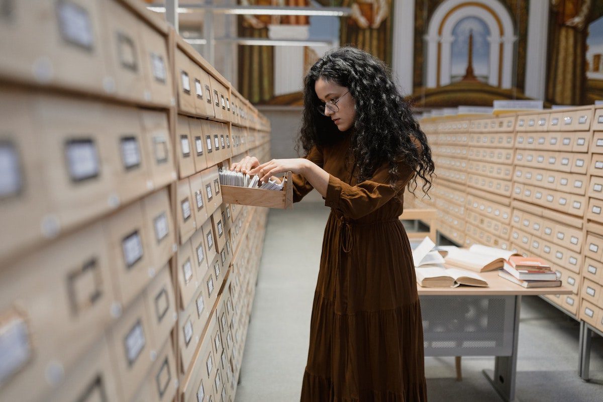 A person in brown dress searches through a Rolodex among other archival shelves Best Online Library Science Masters Degrees