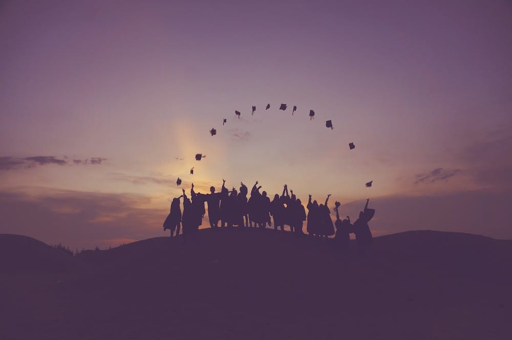 A row of graduates on a hill throwing their hats in a perfect rainbow shape.
