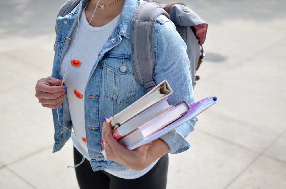 a woman in a blue denim jacket holds her study books and backpack
