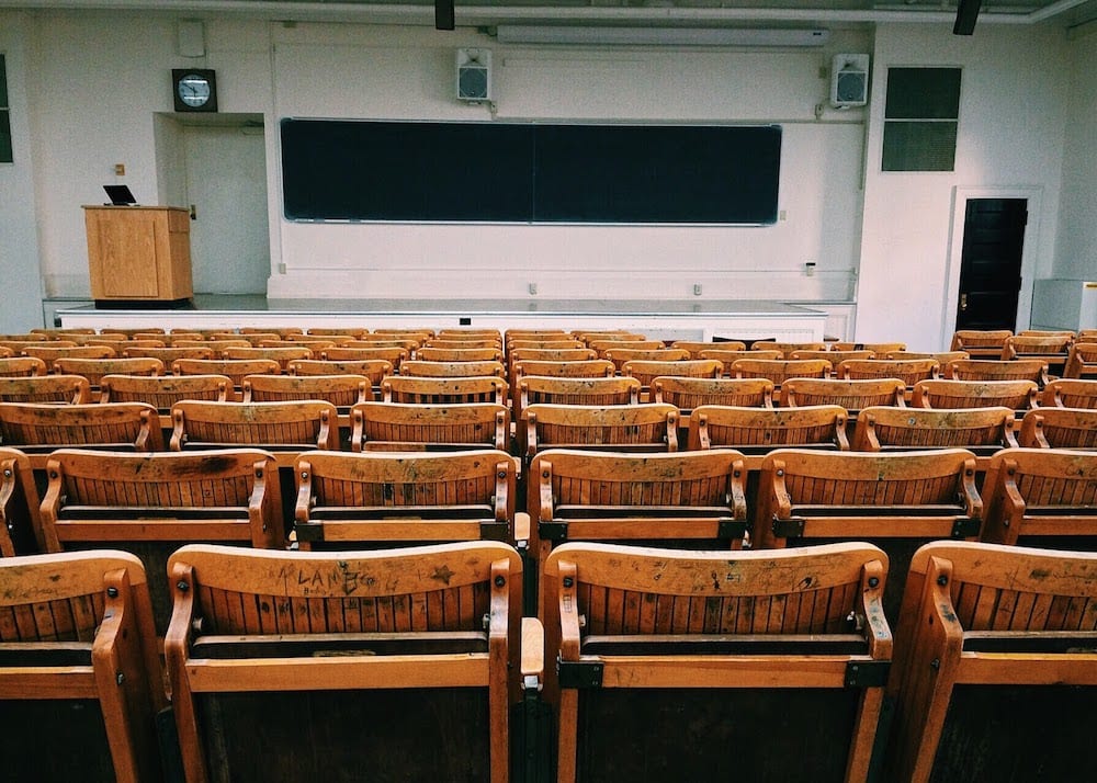 rows of empty wooden university chairs onlook an empty professors podium
