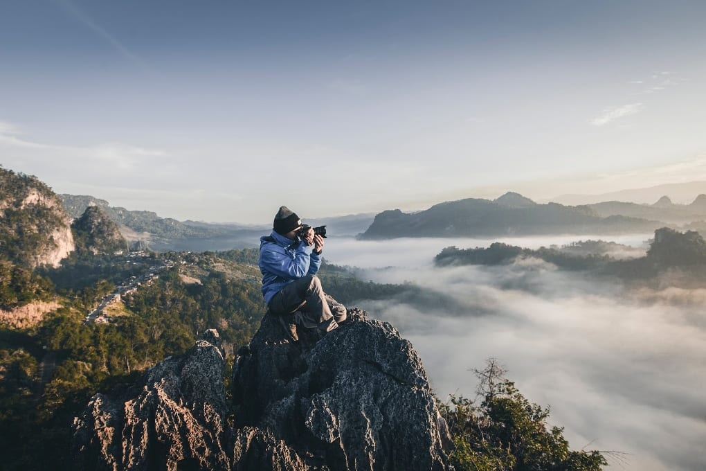 a man sits on a cliff top and photographs a misty mountain landscape