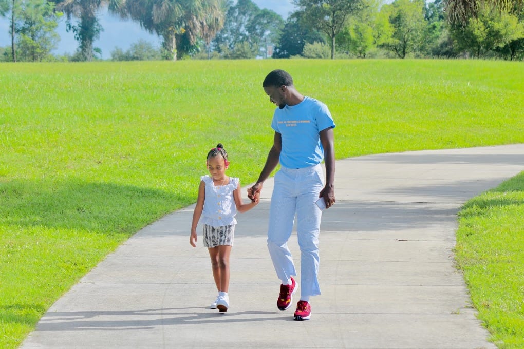 Man in blue t-shirt holding hands with little girl in white shirt and walking on sidewalk at park
