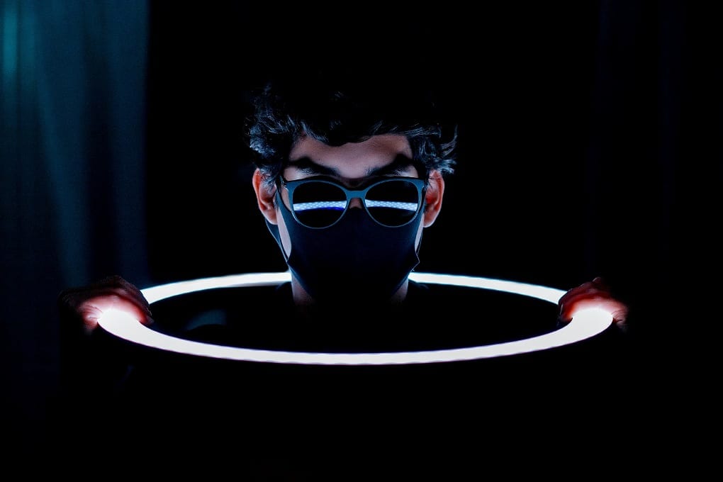 A person in a black mask and sunglasses with an LED ring light around the face.