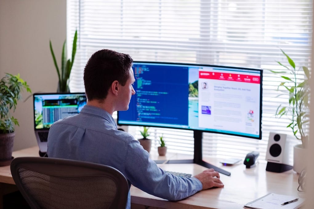 Man in blue dress shirt sitting in front of desktop monitor screen displaying code