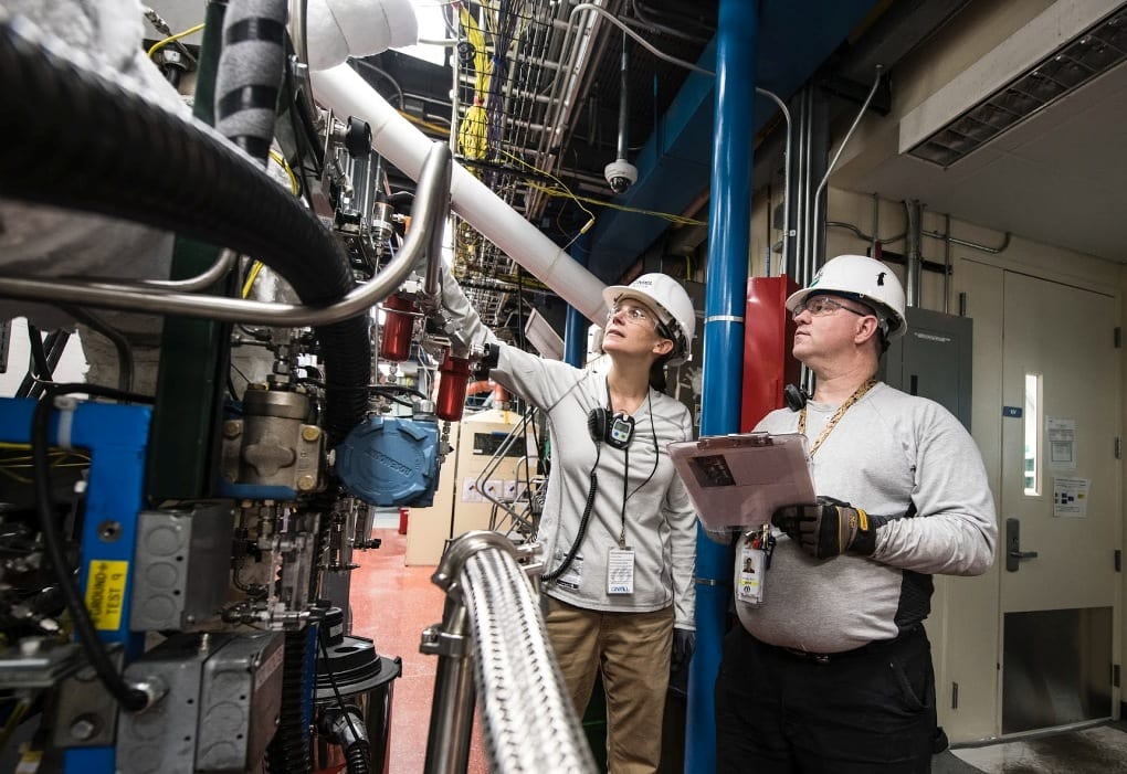 man and woman in grey long-sleeved shirts with white hard hats inspect machinery