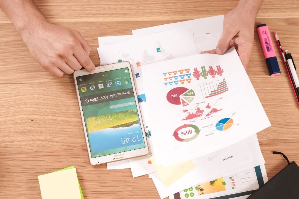 Person holding white paper with colorful graphs over wooden desk next to tablet