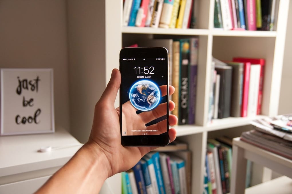 In a room with a large white bookshelf and other white furniture, a person’s hand holds up an iPhone that shows a 3D image of Earth hovering above a digital reconstruction of the part of the hand that is, in reality, behind the iPhone.