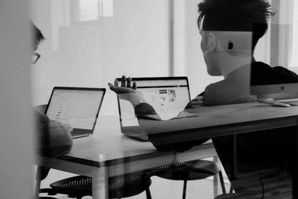 Through the window of a conference room, two people with their backs turned are seen discussing something on their laptop screens.