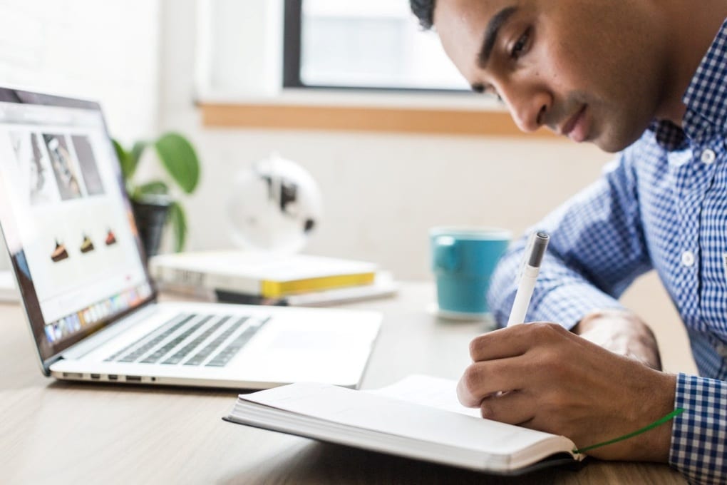 Man wearing blue button-down shirt sits at a desk with a blue coffee mug, model of the earth, and houseplant to his right and a laptop in front of him; resting on the desk below his left shoulder is a notebook, in which he takes notes with his left hand. 