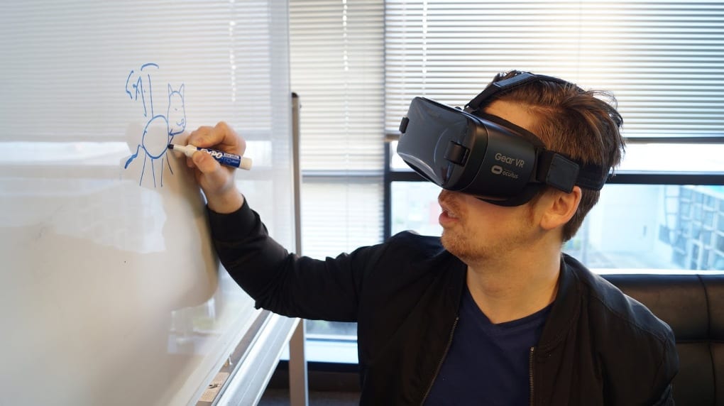 A man wearing a blue shirt, black jacket, and black VR headset draws a four-legged animal on a white board with a blue dry-erase marker. 