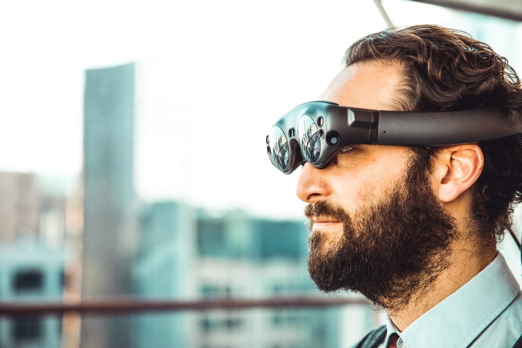 Against a blurry cityscape background, a bearded man wearing a collared shirt, pictured from the shoulders up, looks through a pair of VR goggles.