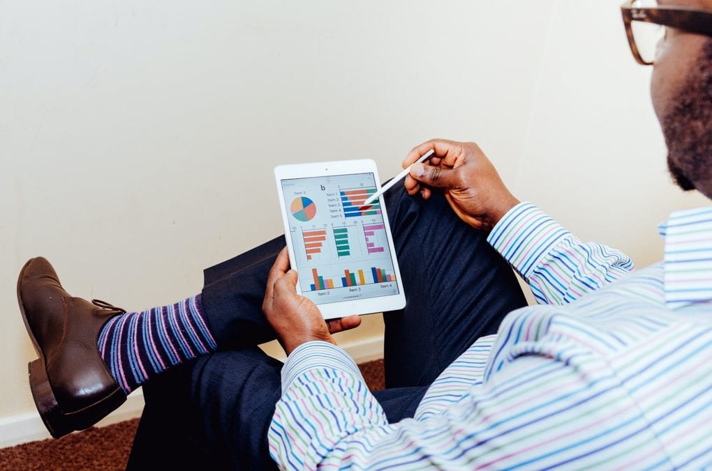 Man in business-casual attire uses a stylus to study graphs and charts on a white tablet. 