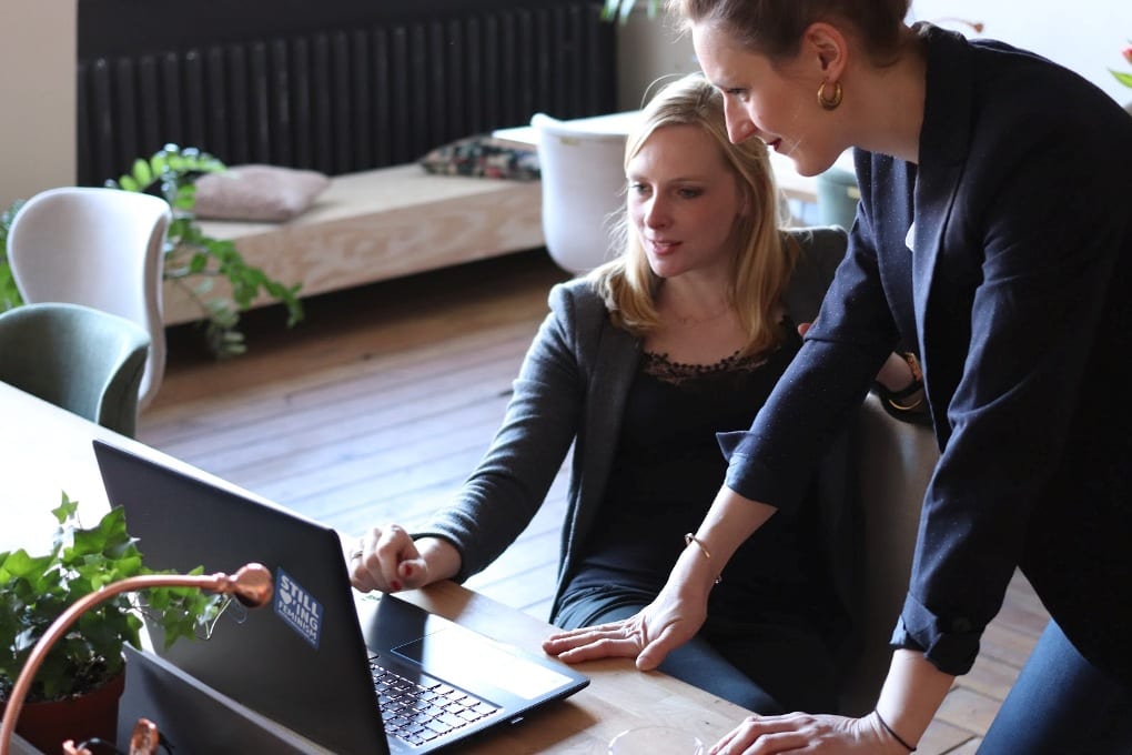 Woman standing over another woman’s desk and helping with laptop