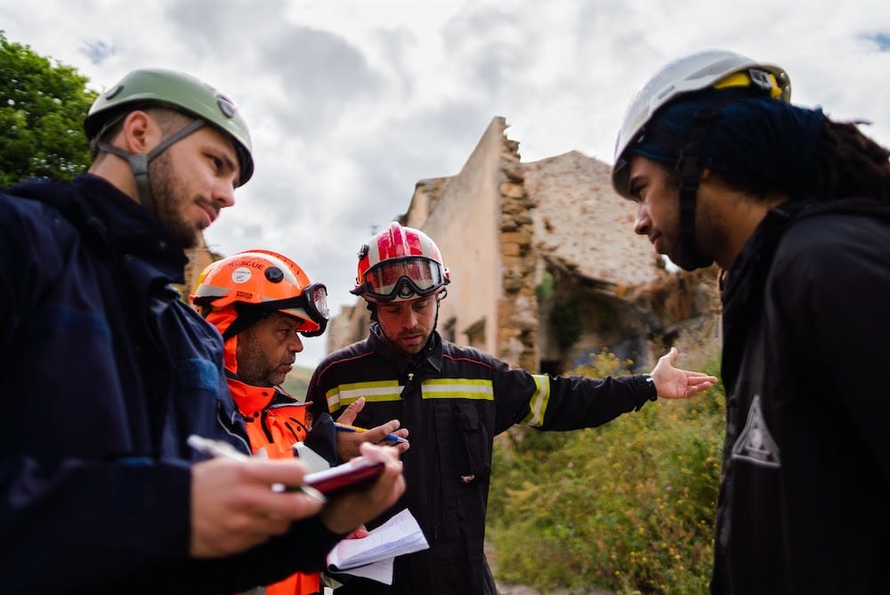 Four men wearing safety jackets and hard hats discuss with one another