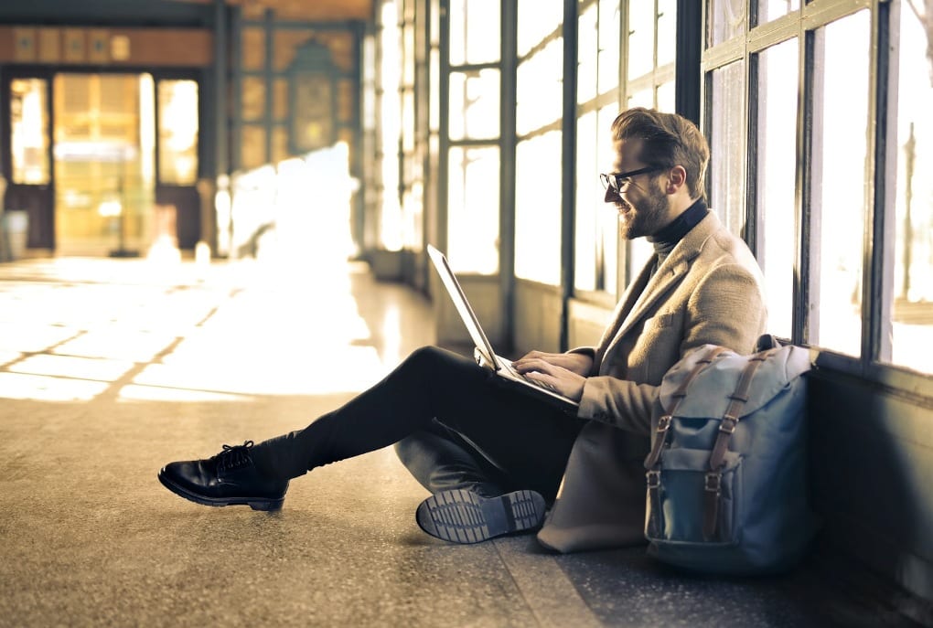 A man in a cream blazer sits on the ground next to his blue backpack and types on a laptop on his lap.