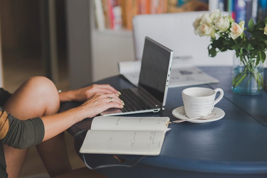 Person typing on their silver laptop with a diary and pen next to them.