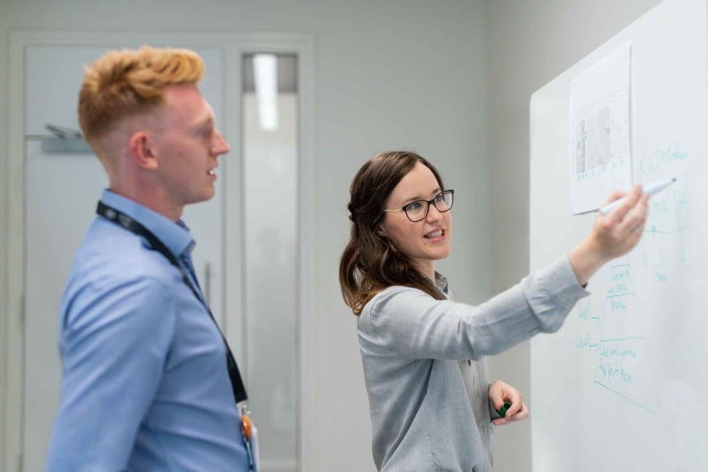 woman holding a dry-erase marker in her right hand explains something to a male colleague, illustrating her point on a white board in front of them