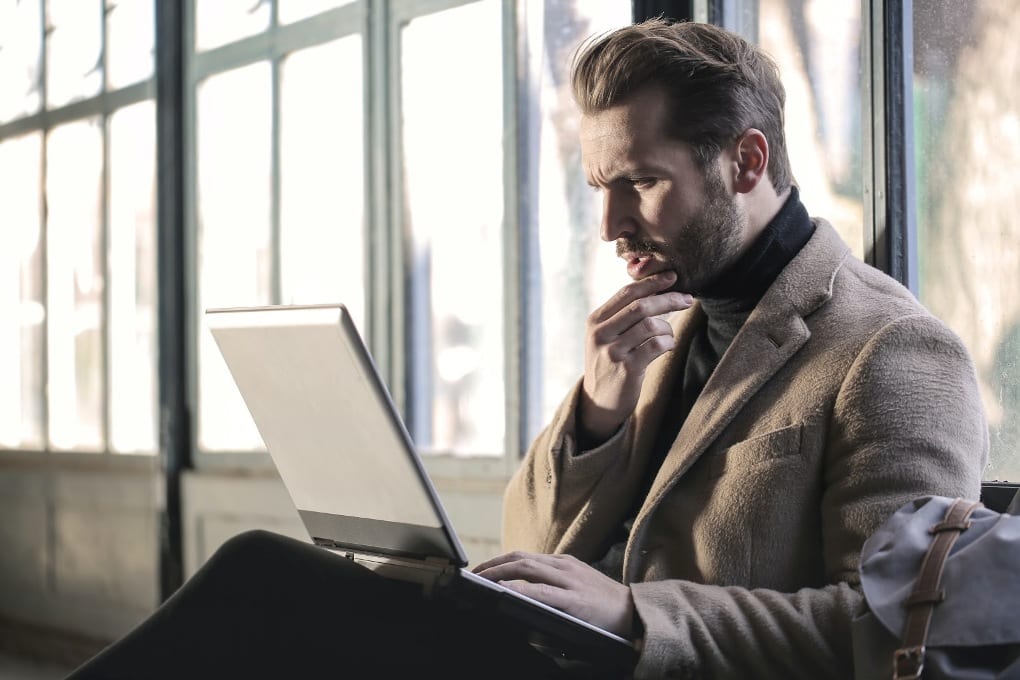 Man in beige jacket holding chin and looking at laptop screen