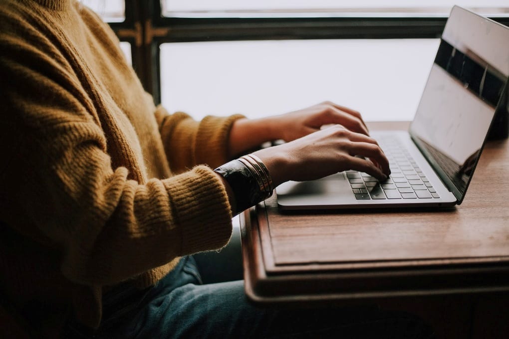 Person in yellow sweater sitting at wooden desk against window and typing on laptop