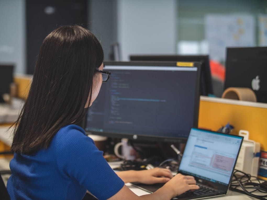 In a computer lab, a woman wearing a blue shirt types on her laptop, with a second monitor displaying a portion of her work in the background.