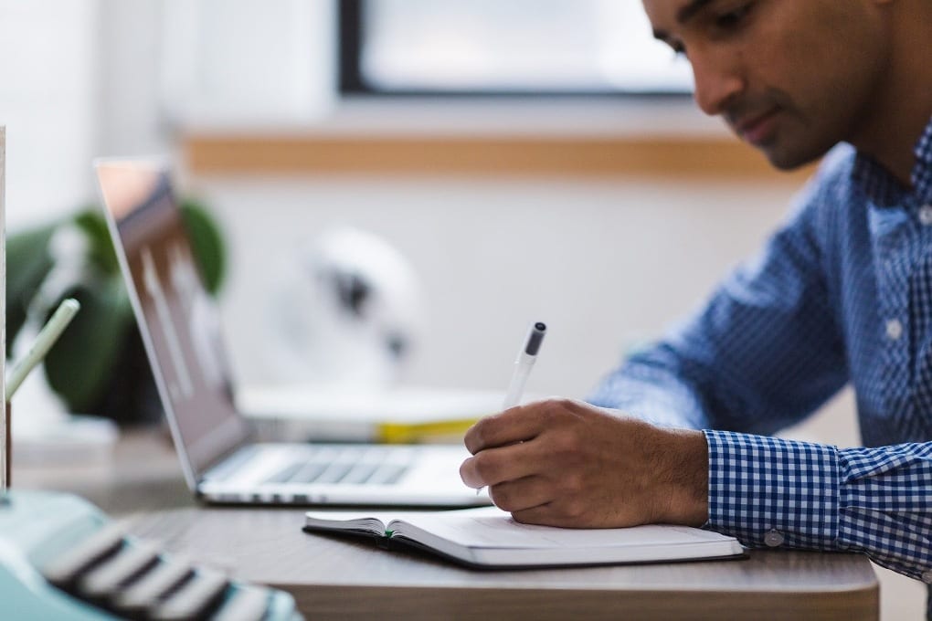 A man sits in front of his laptop, writing in a notebook.