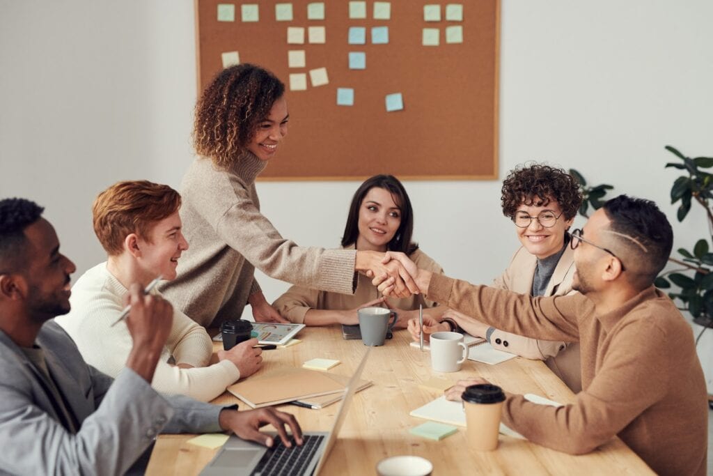 Two people shake hands over a meeting table with six team members sitting around it