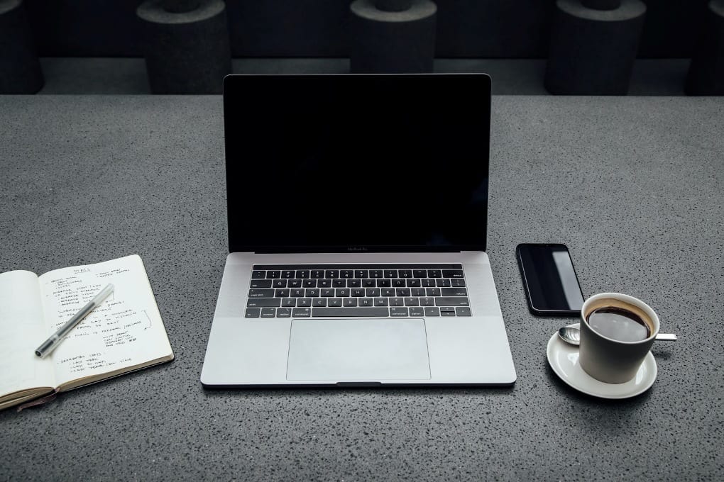 Black and silver laptop on gray desk in between open notebook and coffee mug