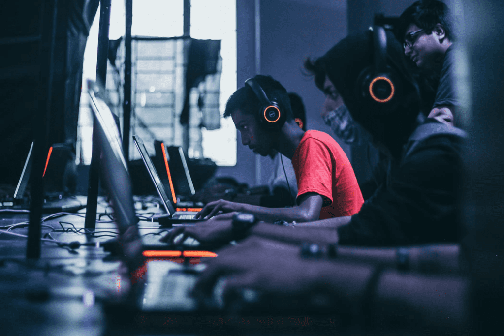 Several young adults sitting in a dark room playing games on laptops and wearing large headphones.