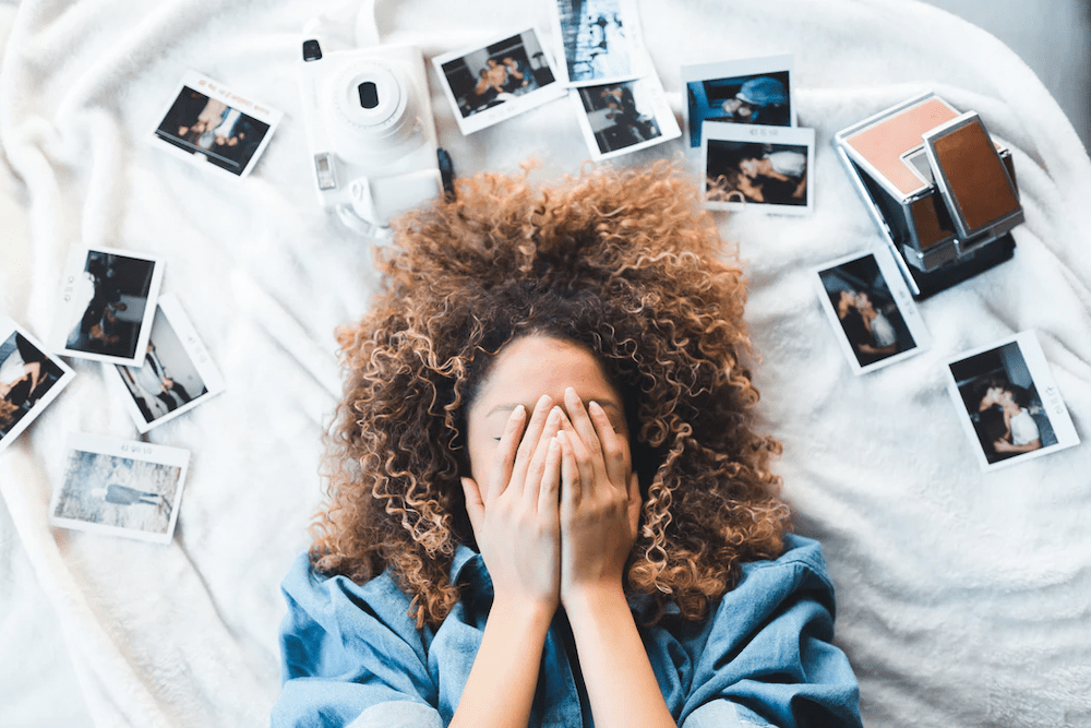 A woman lying on a bed with white covers, a polaroid camera, and various polaroid pictures surrounding her, she is covering her face with both her hands.