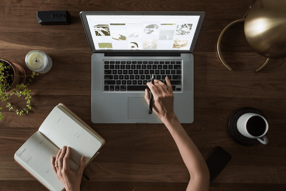 Person’s right hand holding a pen while typing on a laptop, and left hand holding open a notebook.