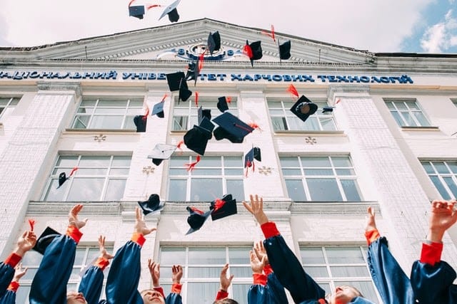 students in graduation robes throwing their hats in front of a white concrete academic building